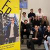 8 ungdommer sitter i ei trapp foran en hvit vegg. En gul plakat  med bilde av en ung gutt og teksten "Vi er ungdommens talerør i Nordland" står ved siden av dem.