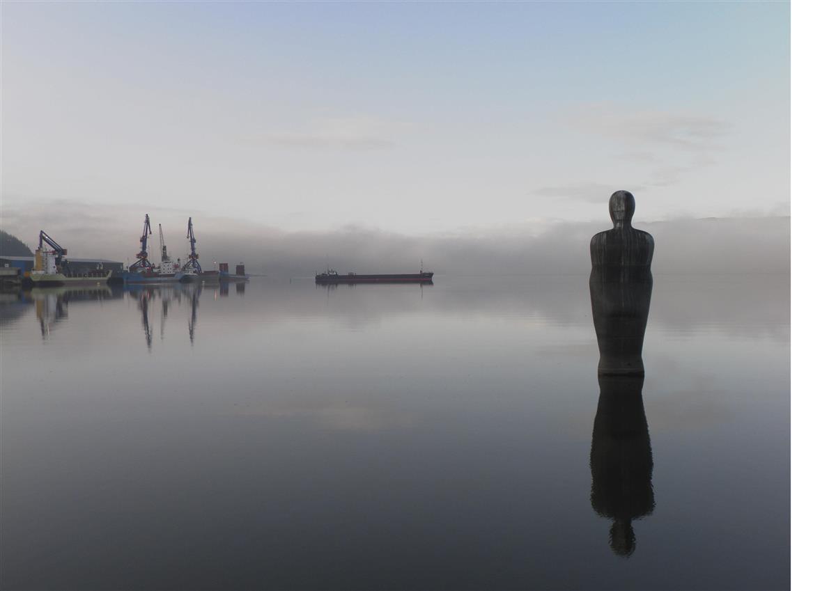 Bilde av skulpturen Havmannen som står ensomt i havet i Mo i Rana.  Skulpturen speiles i vannet. Det er rolig stemning med lavt liggende tåke og fjell i bakgrunnen under en pastell blå himmel. Det er industrianlegg i bakgrunn. - Klikk for stort bilde