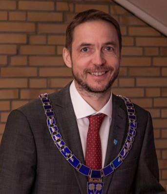 Smilende mann i ordførerkjede, foran mursteinsvegg. - Klikk for stort bilde