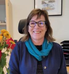 Smilende kvinne med blått skjerf og blå jakke i kontormiljø. - Klikk for stort bilde