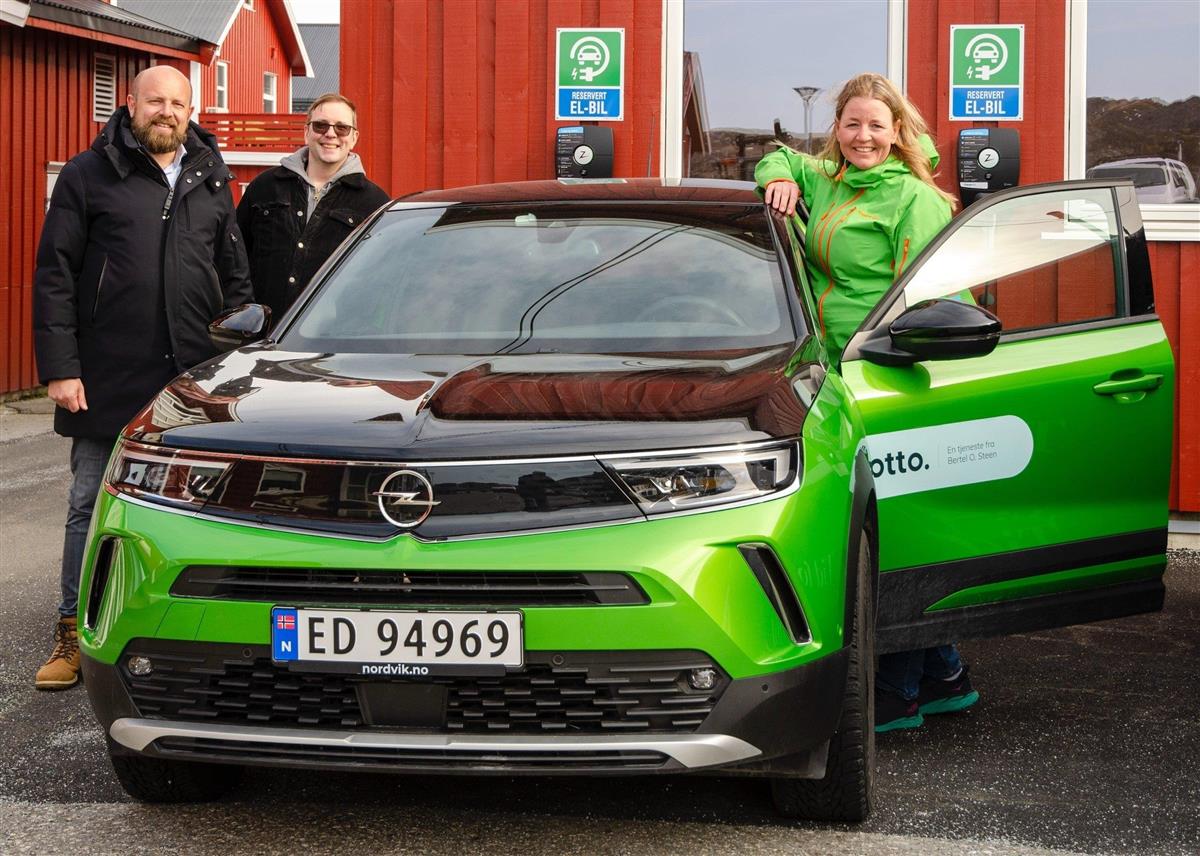 To menn og en smilende dame stående ved en knallgrønn bil. Rødt bygg i bakgrunnen - Klikk for stort bilde