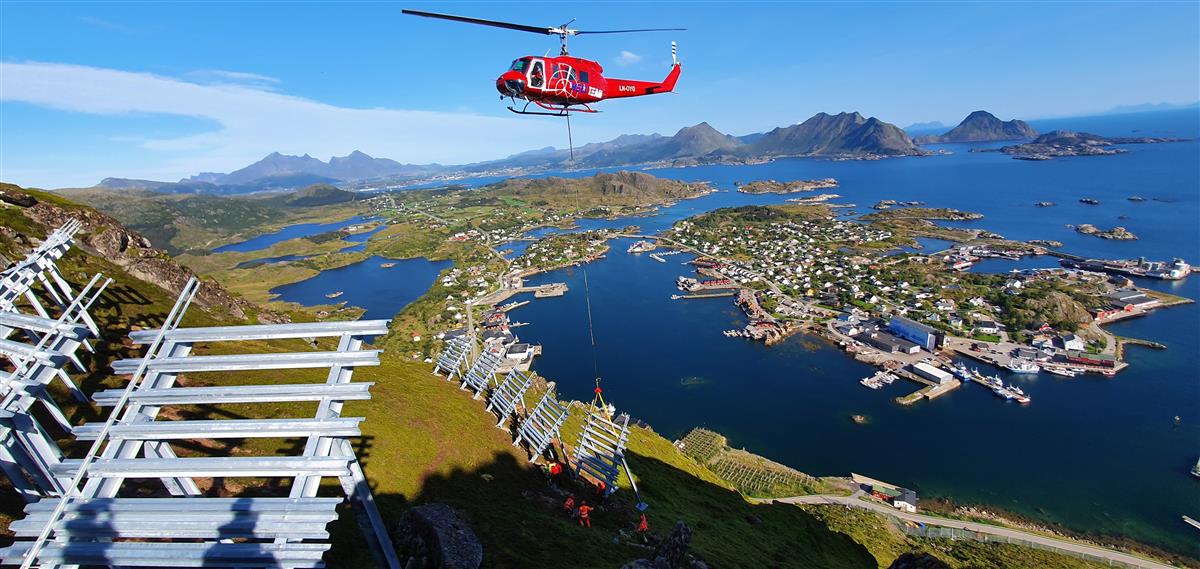 Helikopter svever over rassikringsinstallasjoner i en fjellside, langt over havet. Blå himmel og rolig sjø. - Klikk for stort bilde