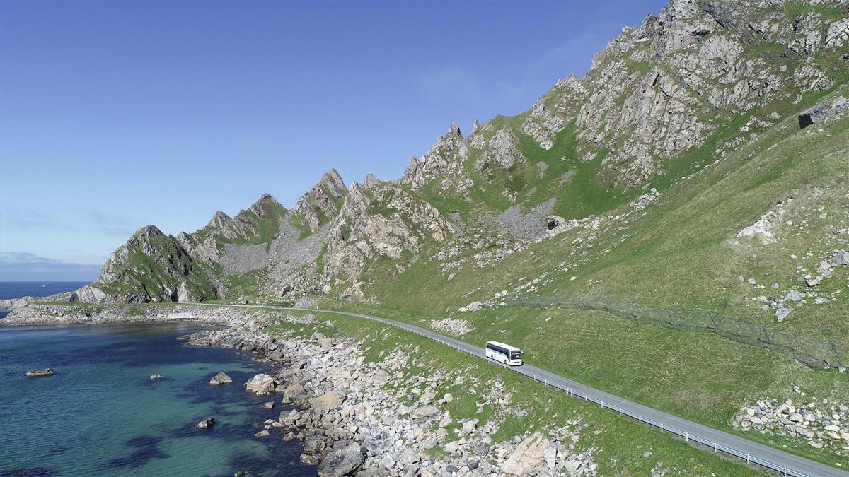 Buss kjører på veg mellom fjell og sjø. - Klikk for stort bilde