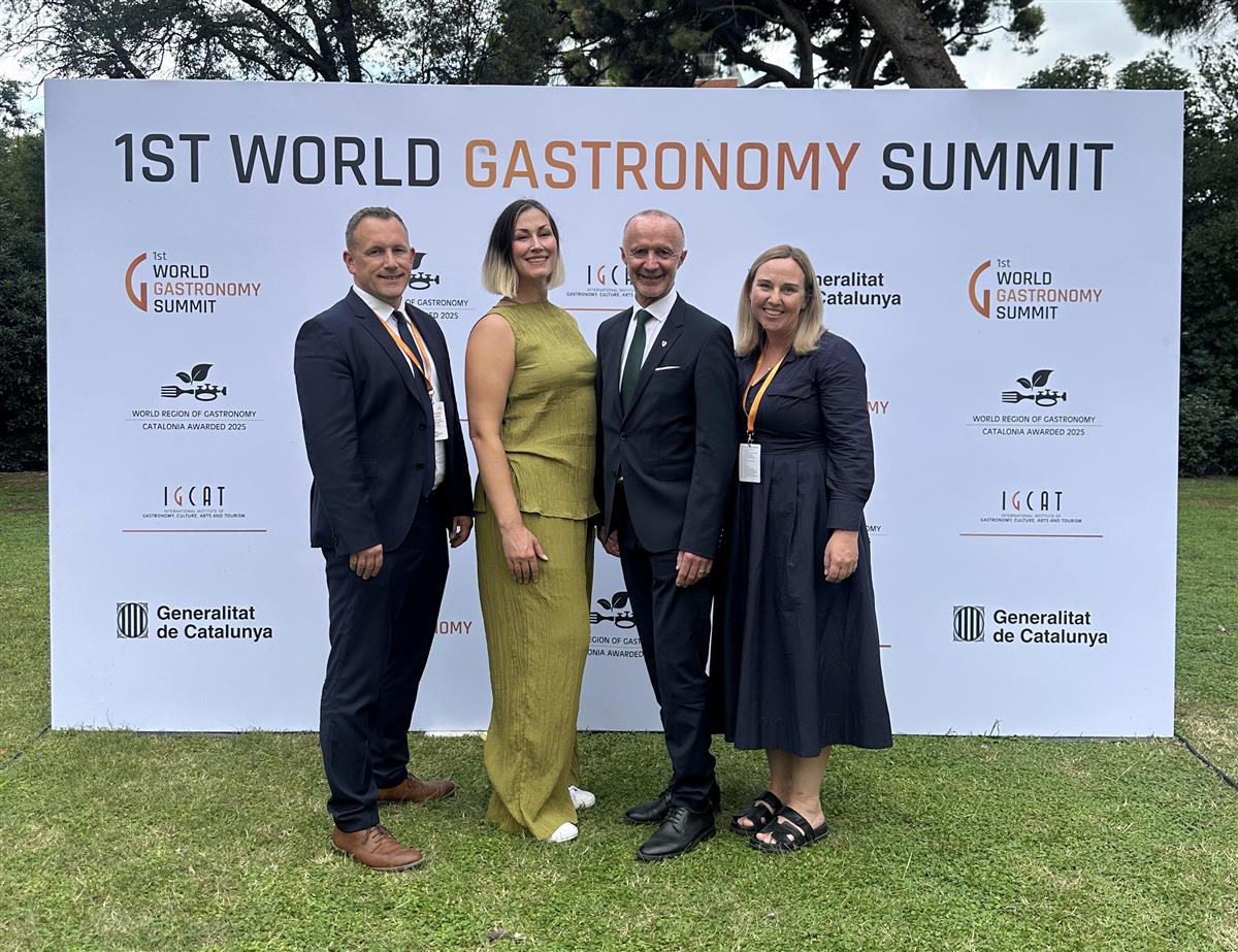 Fire smilende personer fpran et stort skilt. Foto. - Klikk for stort bilde