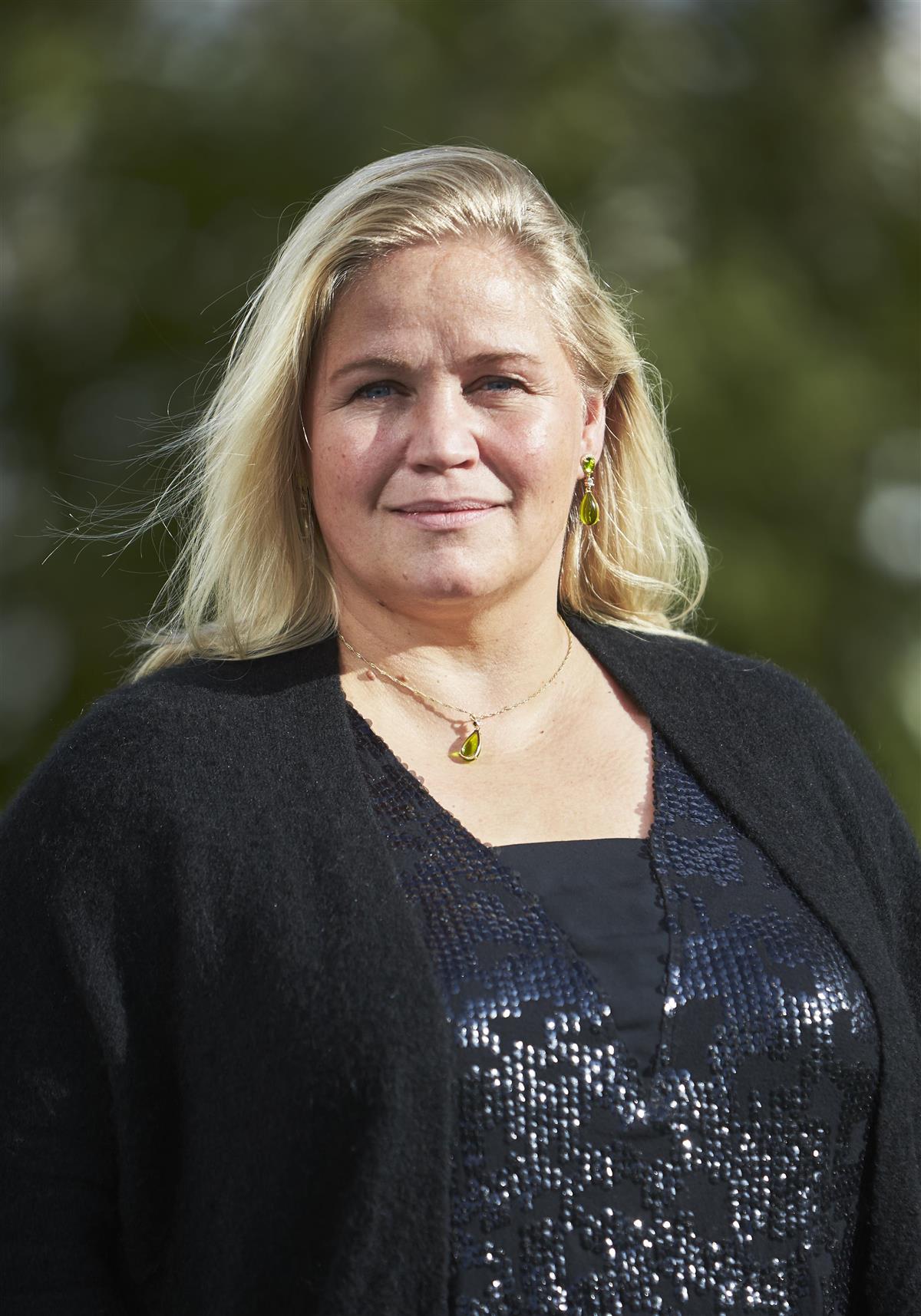 Portrett av blond kvinne med grønne trær i bakgrunnen. - Klikk for stort bilde