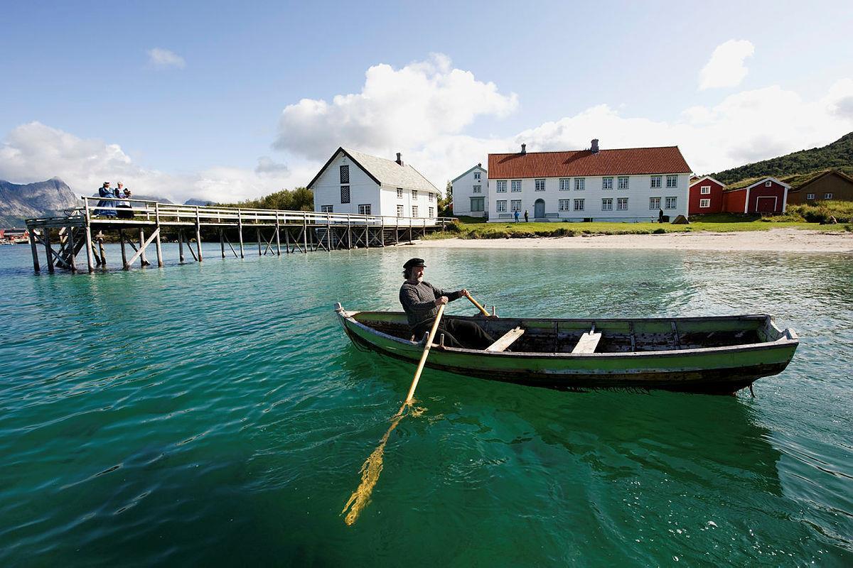 En mann i tradisjonelle klør ror forbi ei havn på Kjerringøy - Klikk for stort bilde