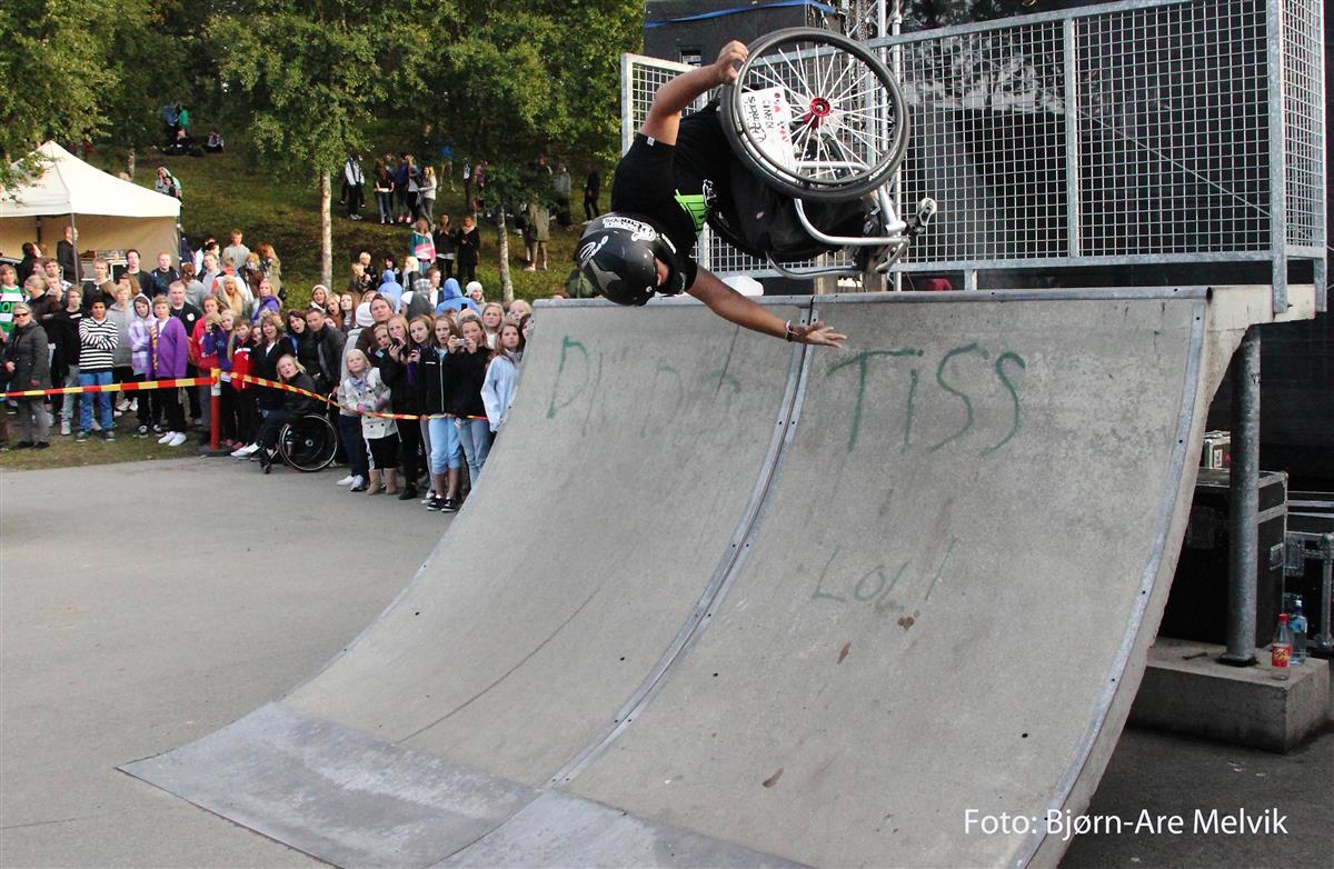 Person i rullestol utfører et spektakulært akrobatisk stunt i en skatepark. Utøveren er opp ned i luften over en rampe mens han holder fast i rullestolen. En stor folkemengde står bak sperrebånd og følger oppvisningen. Arrangementet foregår utendørs med trær og publikum i bakgrunnen. Tekst på bildet: Foto: Bjørn-Are Melvik. - Klikk for stort bilde