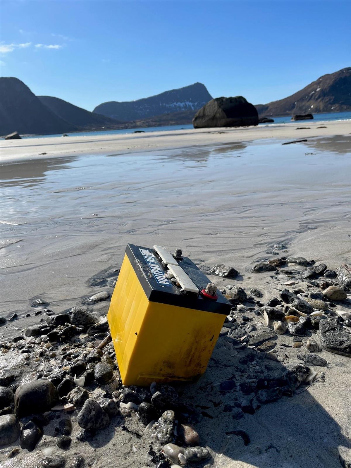 Gult bilbatteri med svart lokk ligger på en hvitsandstrand, med hav og fjell i bakgrunnen. - Klikk for stort bilde