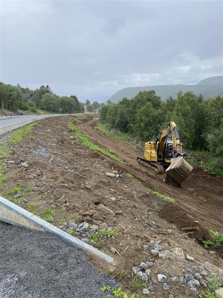 Foto. Skråning nedenfor veg. Gravemaskin i arbeid. Grønne trær - Klikk for stort bilde