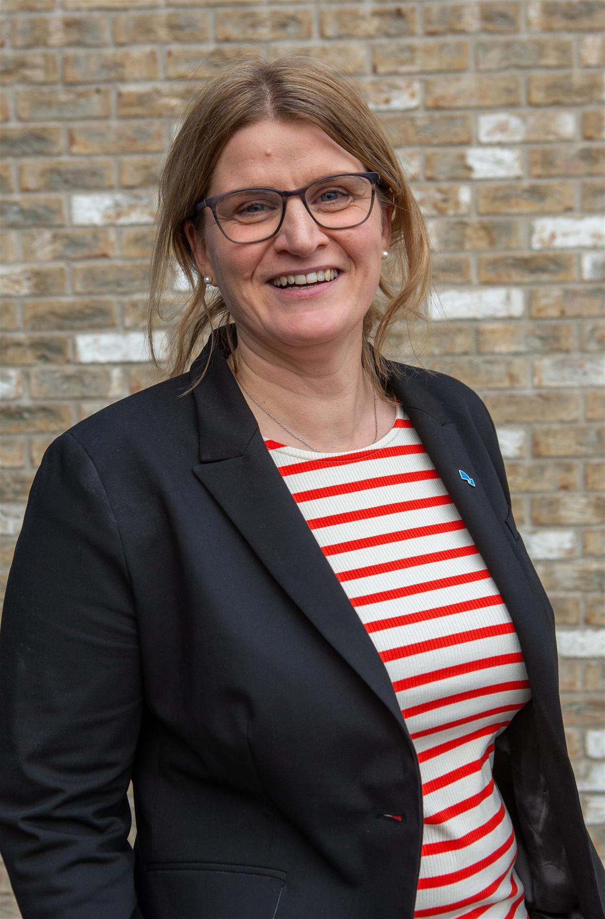 Dame i pen jakke, rød og hvit-stripet genster oppstilt foran murvegg. - Klikk for stort bilde