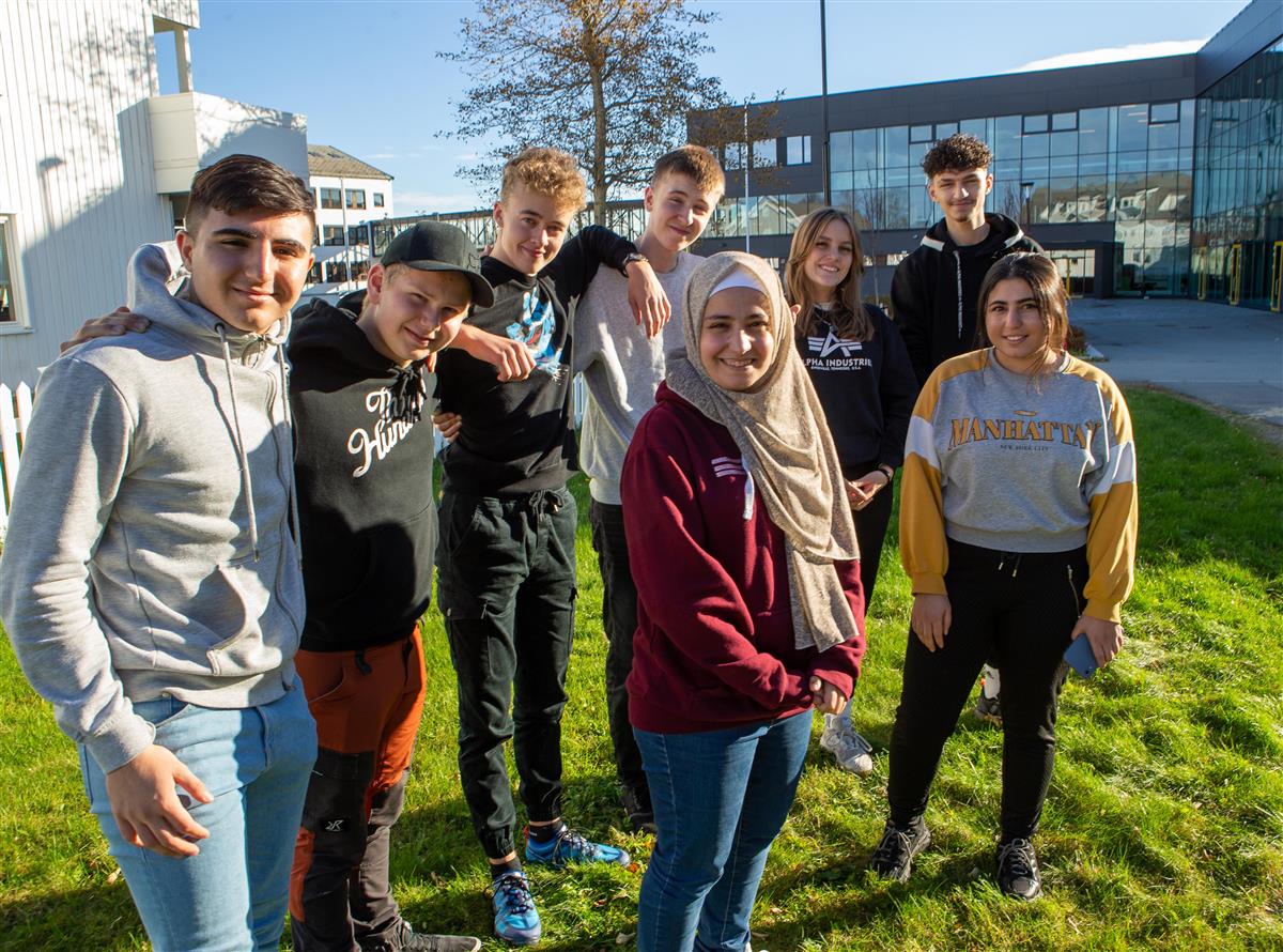 8 blide ungdommer står på en gressplen foran et skolebygg. Foto - Klikk for stort bilde