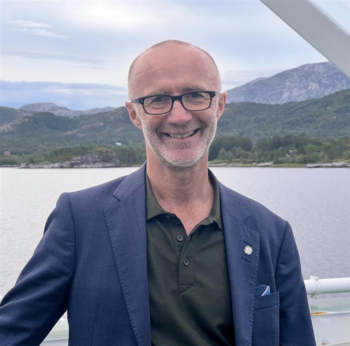 Smilende mann på båt med hav og fjell i bakgrunnen. Foto. - Klikk for stort bilde