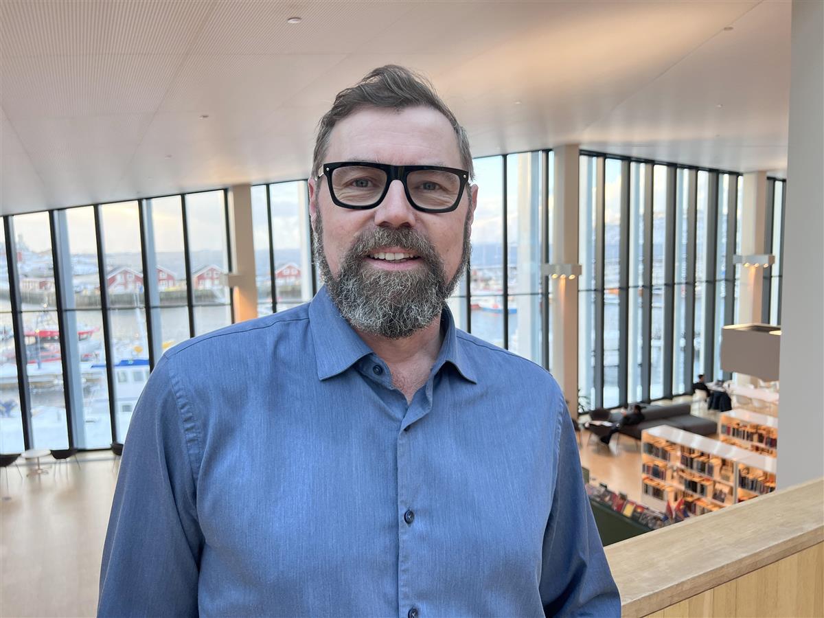 smilende mann inne på bibliotek. Foto - Klikk for stort bilde