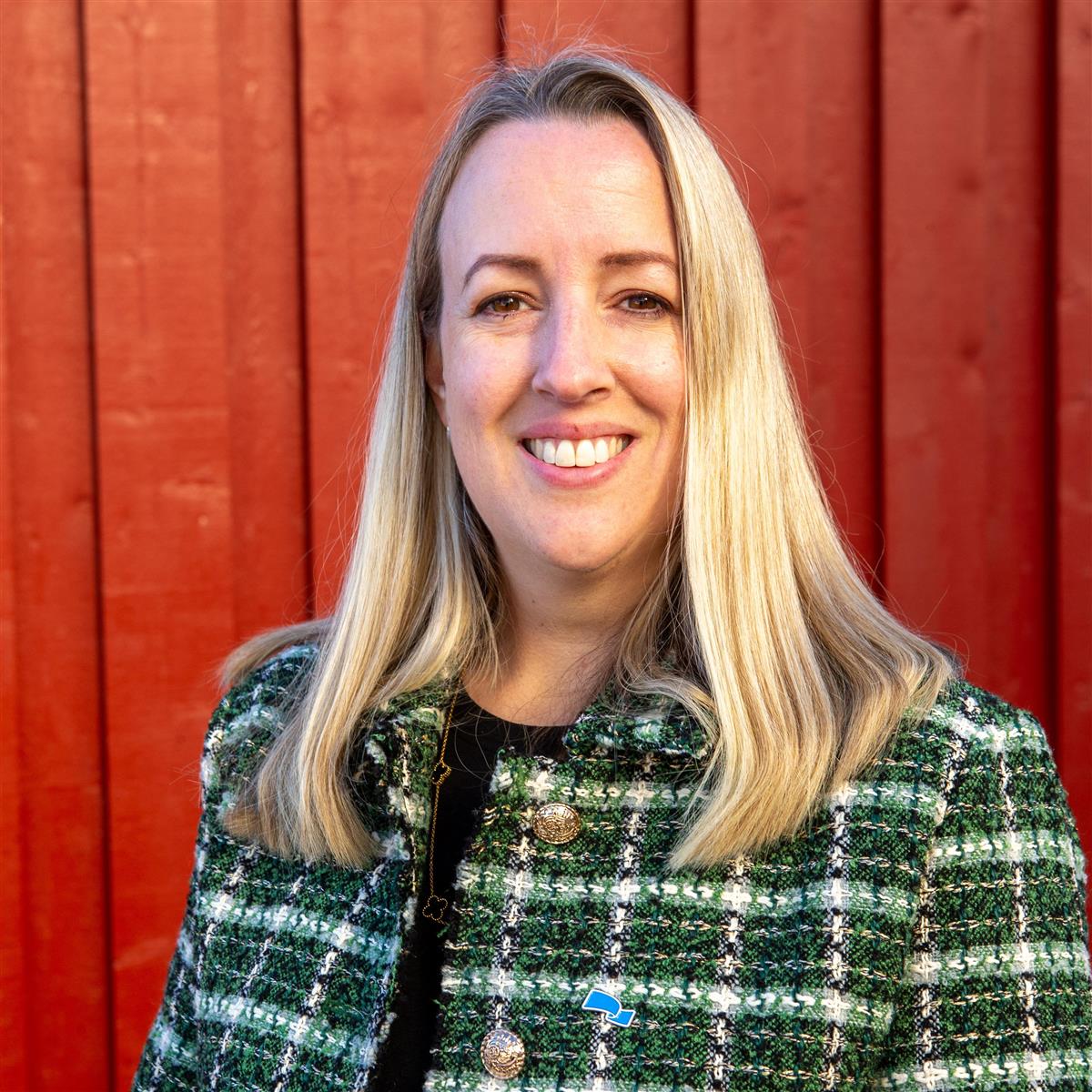 Portrett av blond kvinne i grønn, rutet jakke foran rød vegg. - Klikk for stort bilde