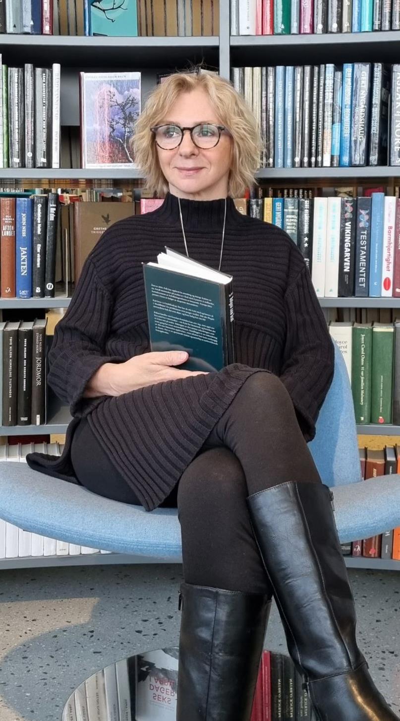 Kvinner sittende i lyseblå stol med en bok i hånden. Bake henne er bibliotekbokhyller. Foto - Klikk for stort bilde