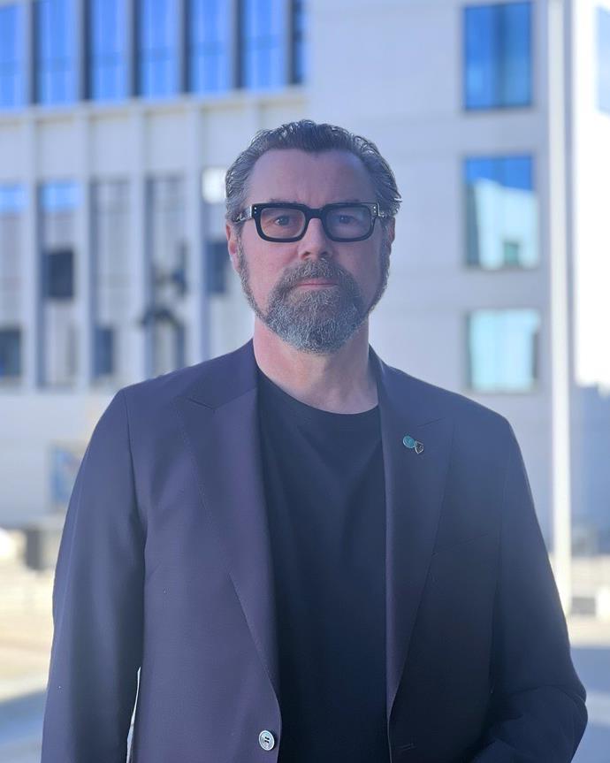 En mann med briller og kort, mørkt hår med grått skjegg står utendørs foran en moderne bygning. Han har på seg en mørk blazer over en svart t-skjorte og ser rett mot kamera med et nøytralt uttrykk.