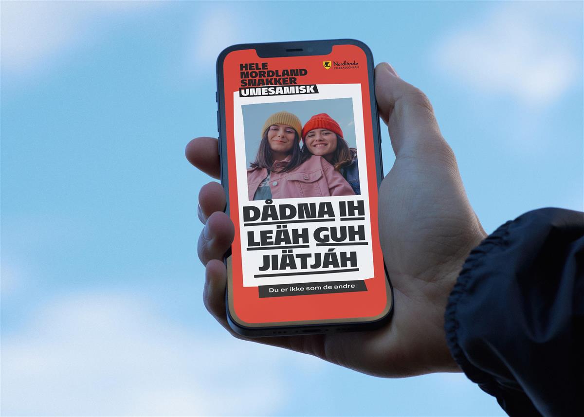 hånd som holder mobil opp mot himmelen. På skjermen er det bilde av to jenter og en tekst på samisk. Foto - Klikk for stort bilde