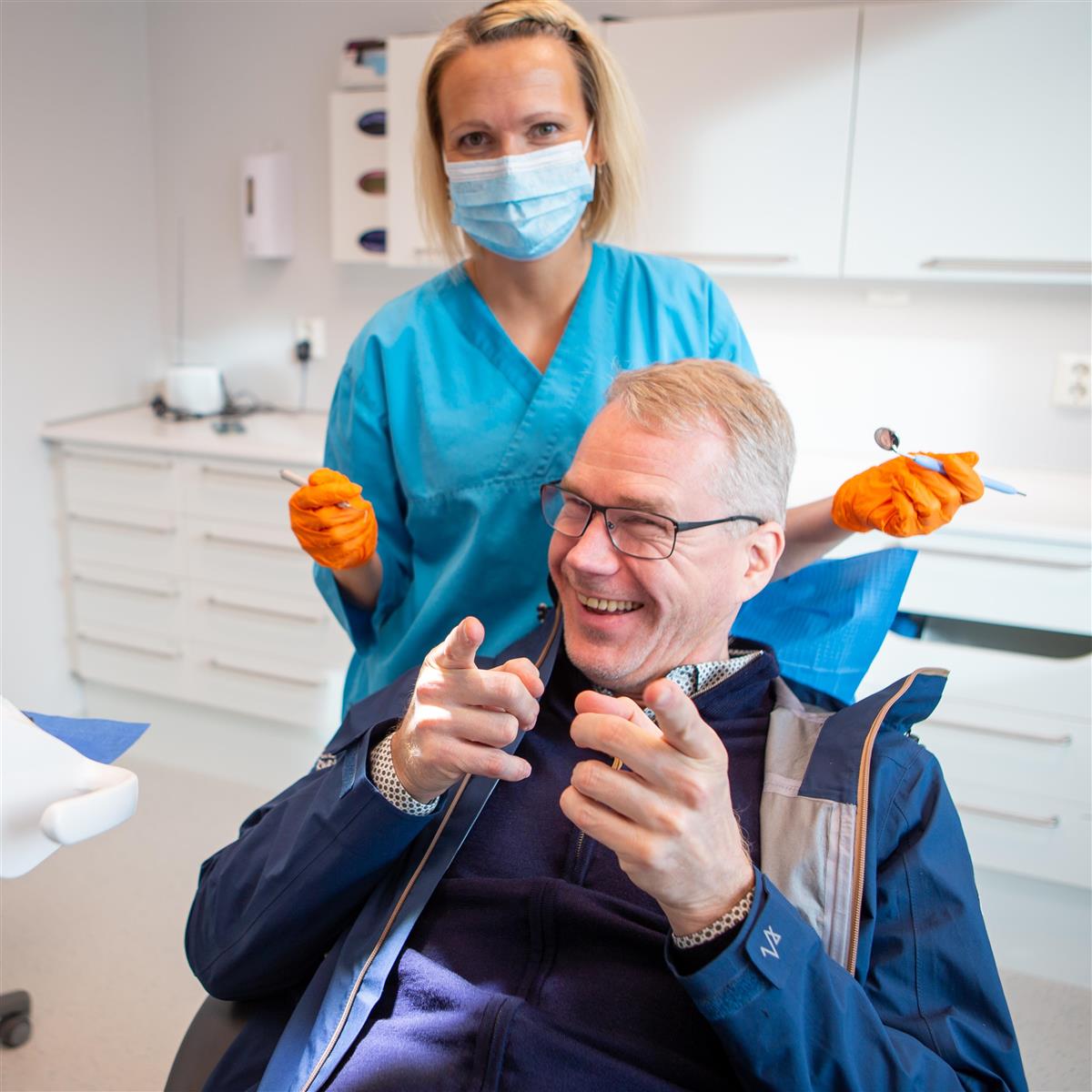 Mann med briller sitter i tannlegestol med et stort smil og med to pekefingre som peker mot fotografen. Over ham står en kvinnelig tannlege med oransje plasthansker og tannhelseutstyr i hendene. - Klikk for stort bilde