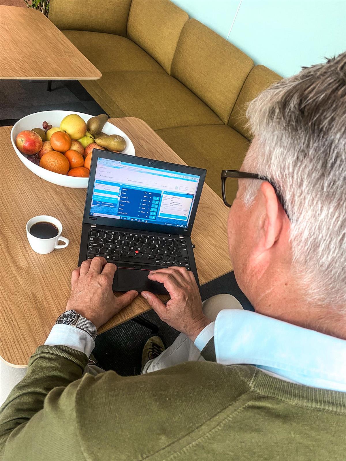 Mann med grått hår og briller sees bakfra mens han jobber på bærbar PC på et stuebord. Han har kaffikoppen til venstre for PC-en og en fruktskål bak. - Klikk for stort bilde