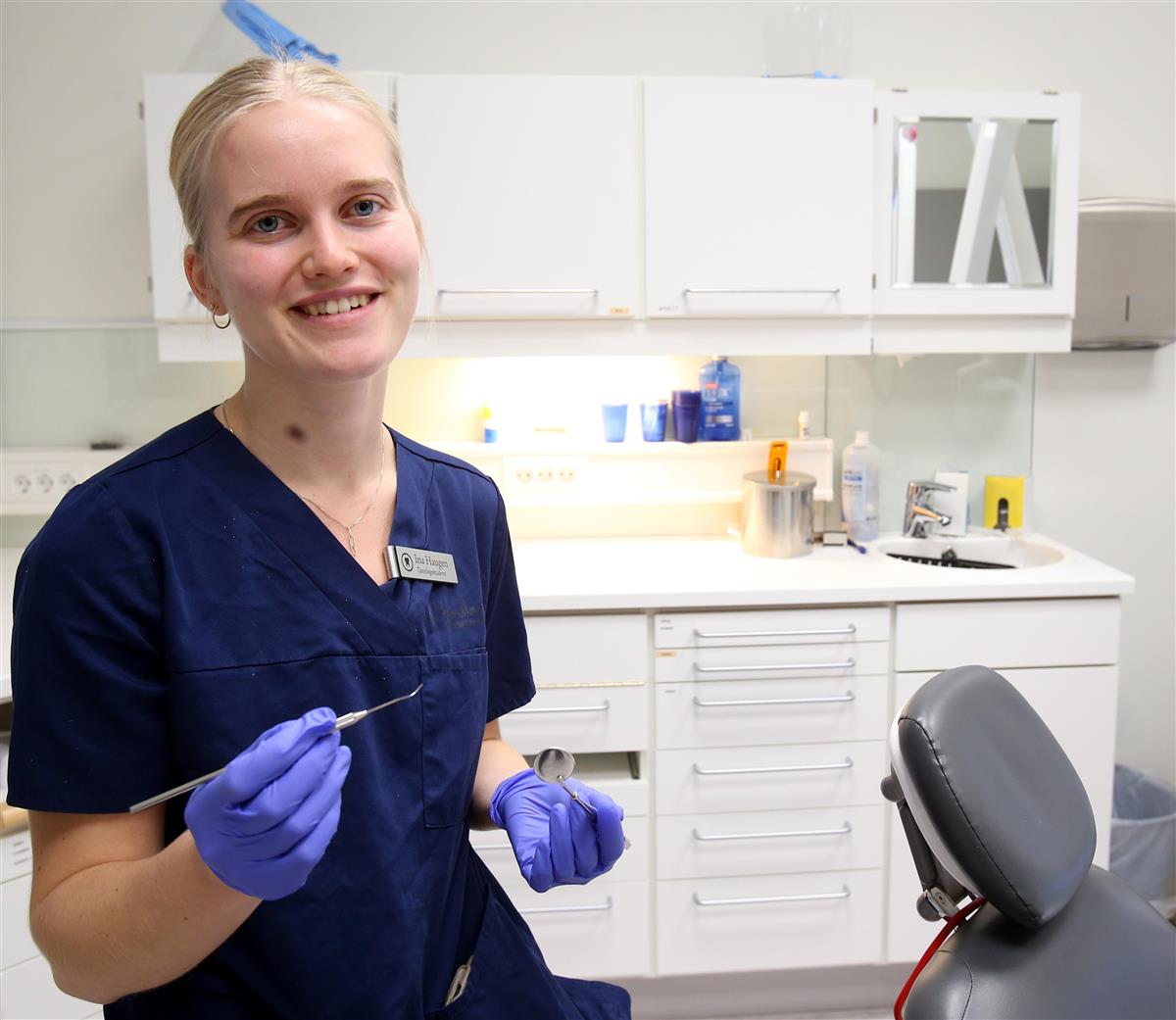 Smilende tannlegestudent i blå arbeidsuniform og hansker sitter i et moderne behandlingsrom og holder tannlegeinstrumenter i hendene. Foto - Klikk for stort bilde