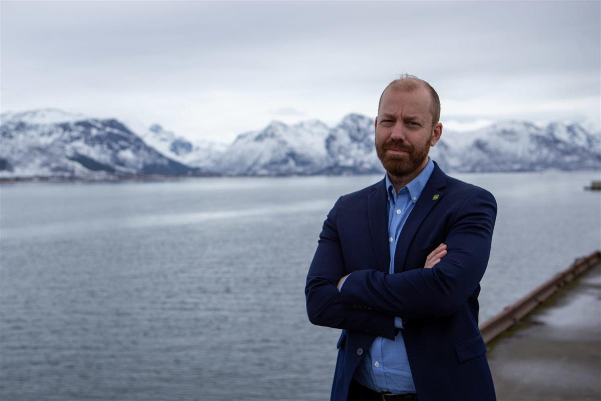 Mann med armene i kryss, hav og fjell i bakgrunnen. - Klikk for stort bilde