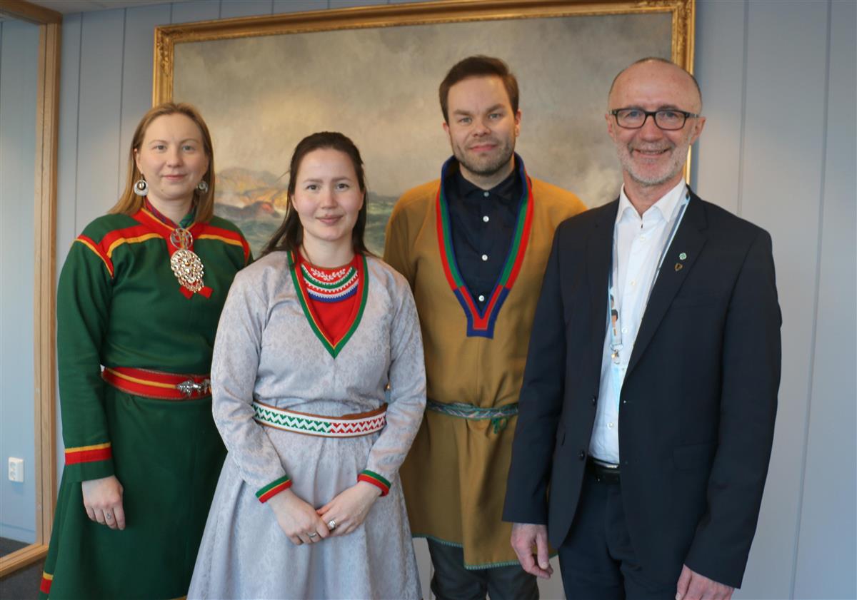 Fire personer oppstilt, tre kledd i samiske kofter og en kledd i dress. - Klikk for stort bilde